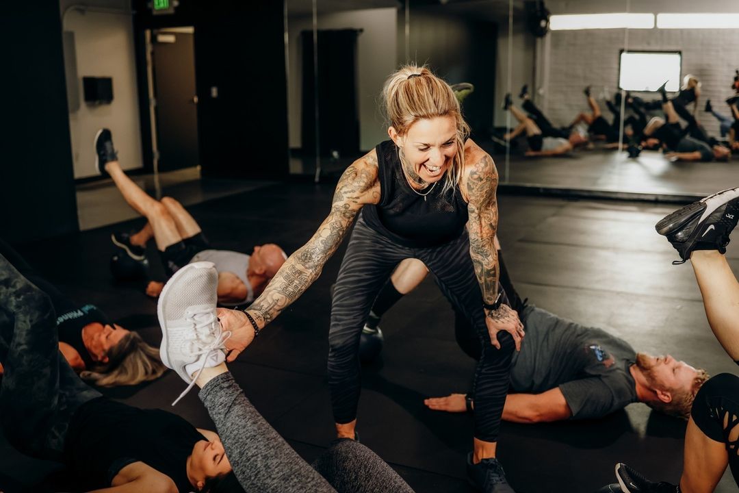 The image shows a lively gym scene with a tattooed female trainer (Carina Reid) enthusiastically engaging with participants during a workout. She’s holding one participant’s leg while smiling broadly, radiating energy and encouragement. In the background, several people are lying on the floor performing leg-raise exercises, reflected in the mirrored wall. The atmosphere feels intense yet fun, with a strong sense of motivation and camaraderie.