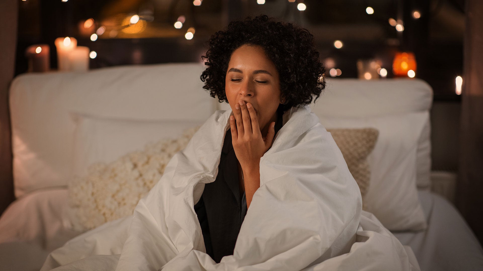 Woman wrapped in a blanket yawning in a cozy room with candles and string lights.