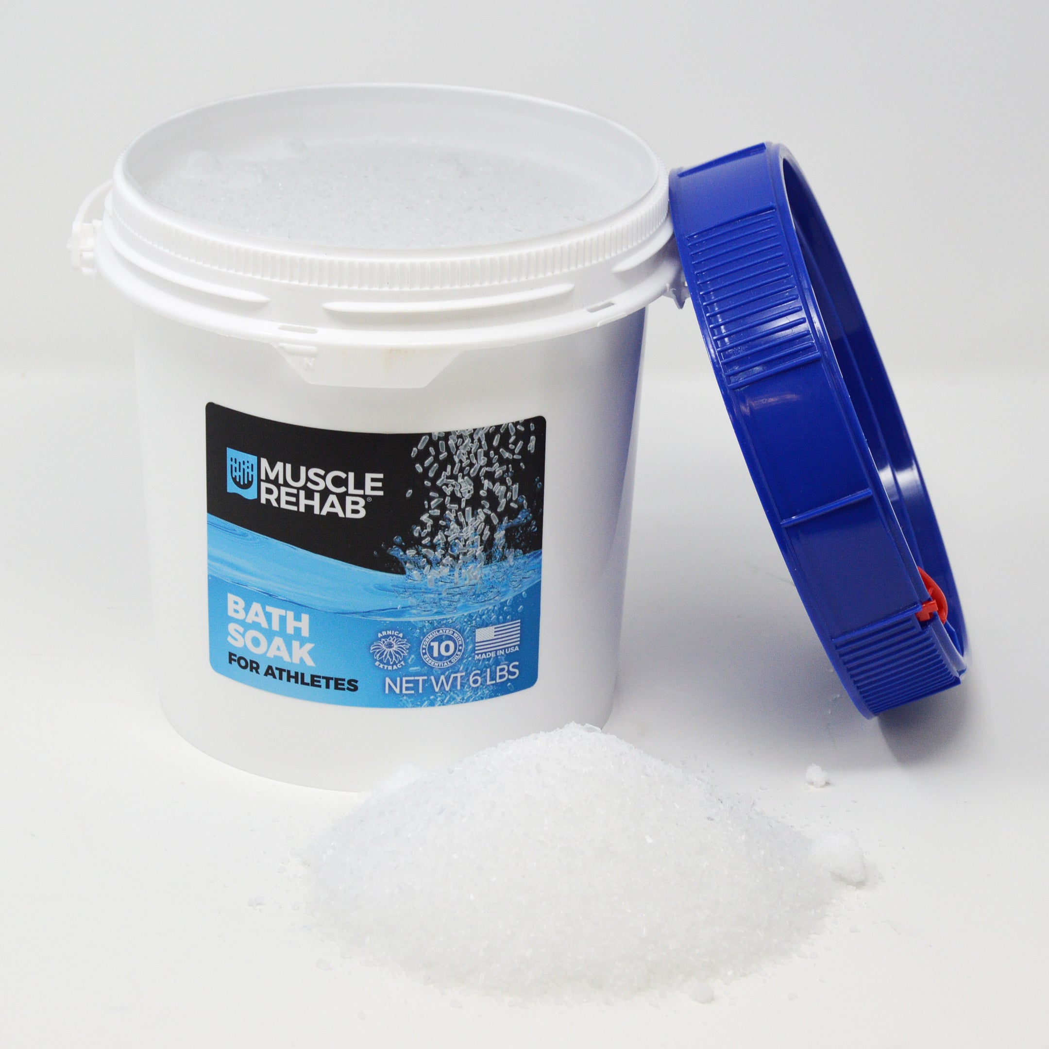 This image shows the Muscle Rehab® Bath Soak (6 lb pail) in a lifestyle/product display:

Product: White pail with a blue lid (lid is removed and placed to the side), showing the front label clearly.

Label Details: Brand logo, “Bath Soak for Athletes” name, “Formulated with 10 Essential Oils” badge, “Made in USA,” and net weight.

Lifestyle Element: A small mound of bath soak granules is poured in front of the container, showing the texture and crystalline appearance of the product.