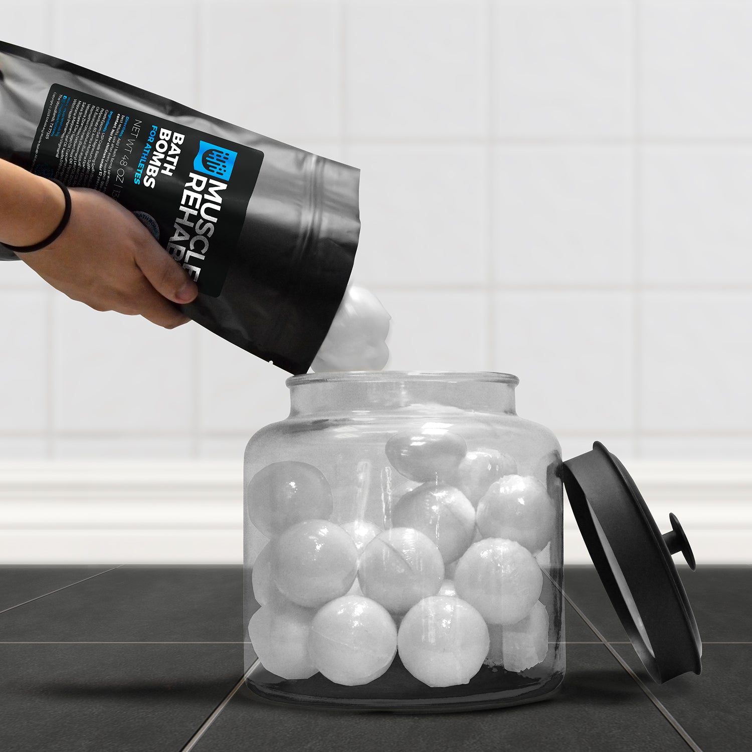 Muscle Rehab bath bombs pouring out from the 12-pack pouch and into a glass container resting on a steamy bathroom floor